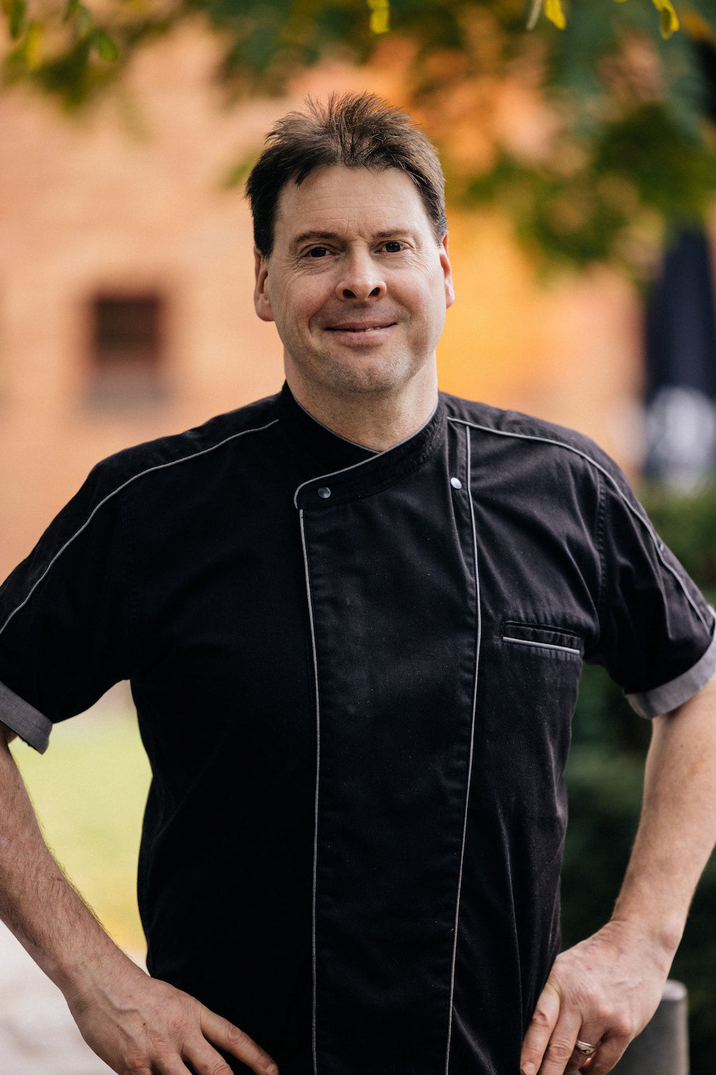Man in a chef's jacket smiling outdoors near a brick building