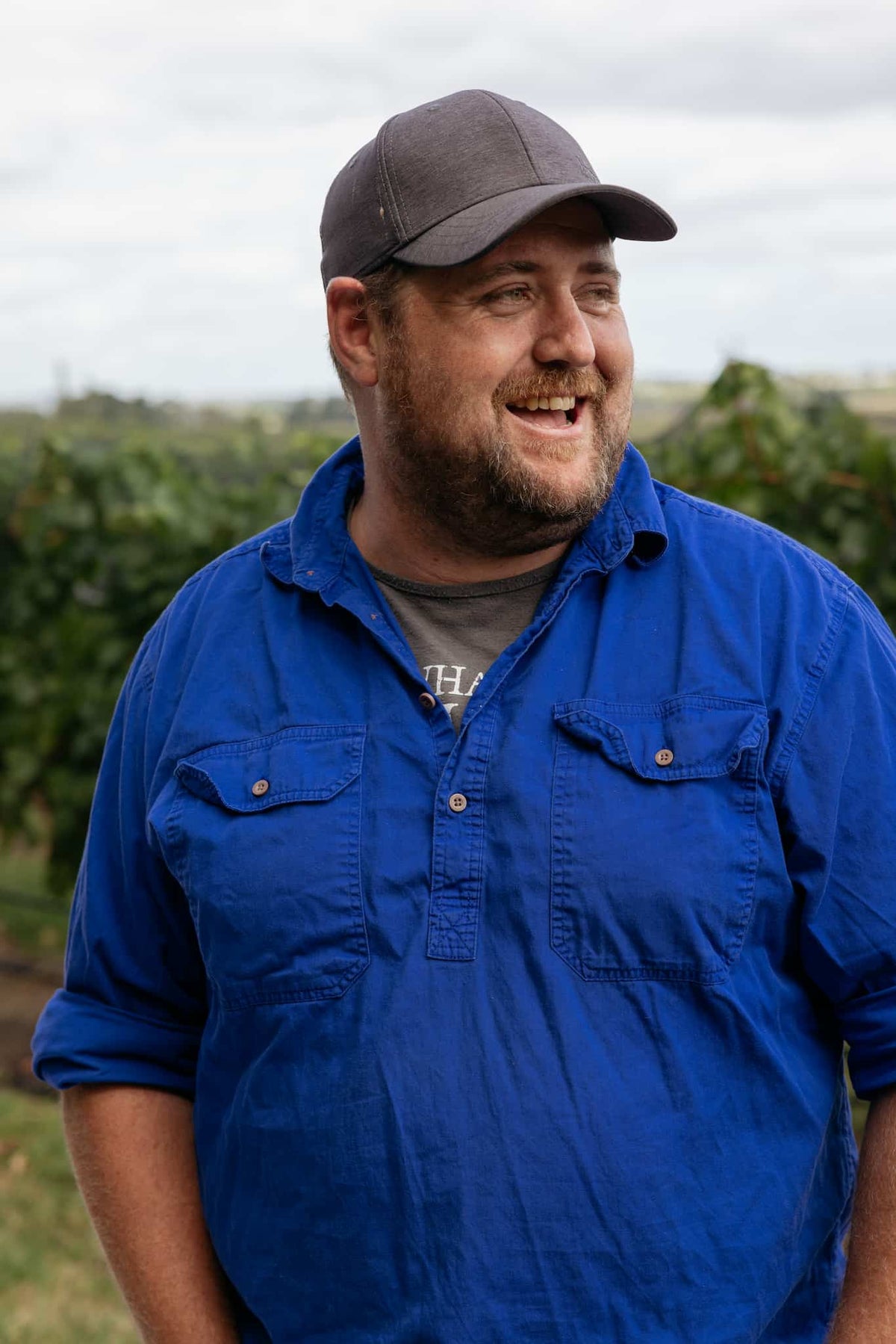 Man wearing a cap and blue work shirt smiling in front of vineyard rows