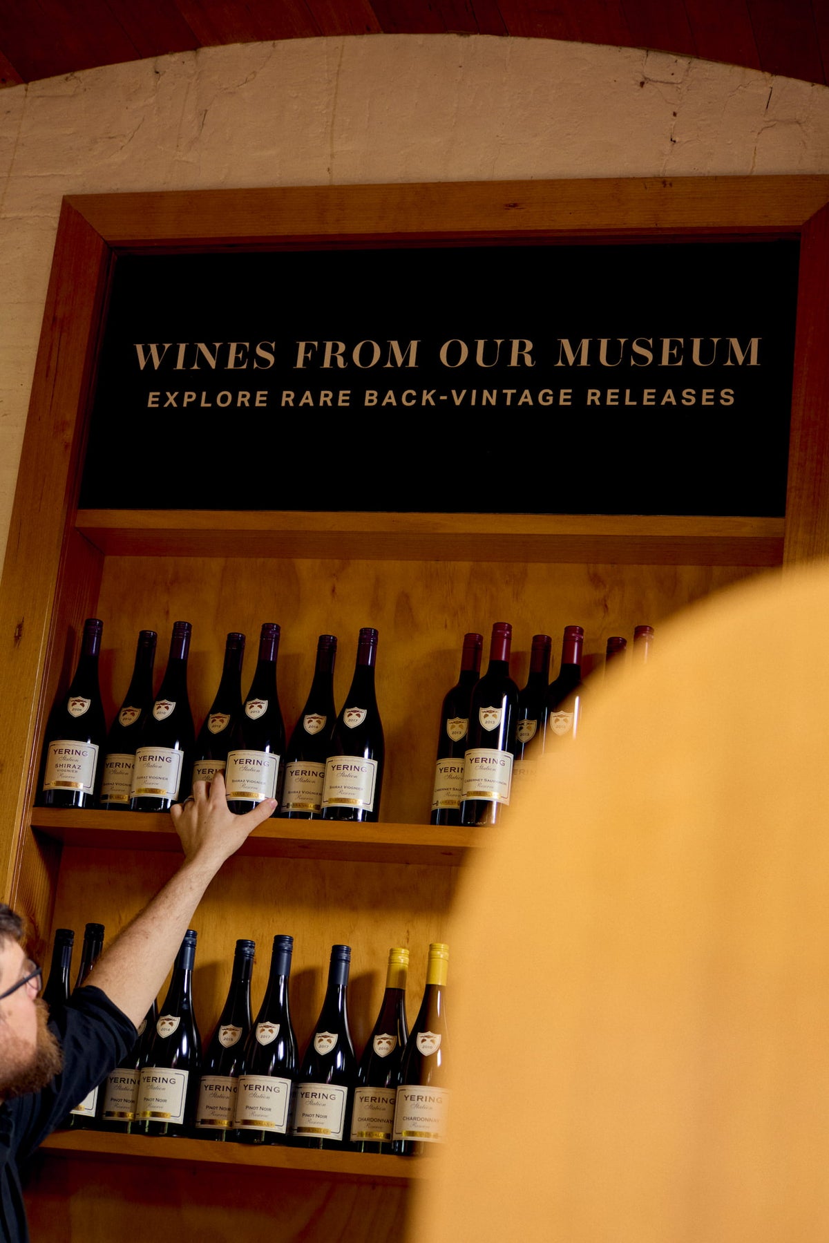 Cellar staff member reaching for a bottle on a wooden display shelf labelled Wines From Our Museum