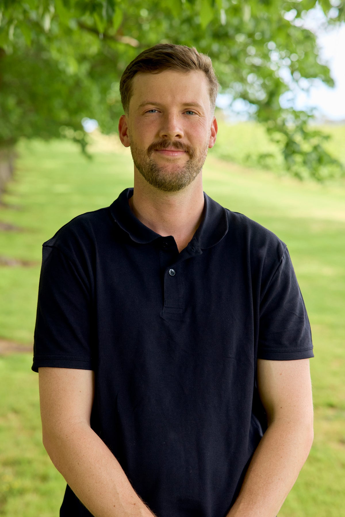Portrait of a man in a black polo shirt smiling outdoors with trees in the background