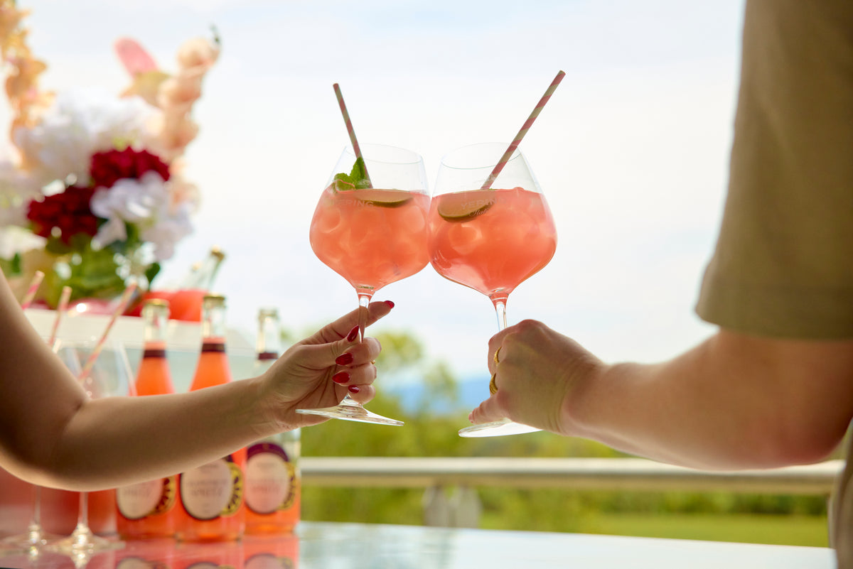 Two people toasting pink spritz cocktails on an outdoor terrace overlooking the vineyard