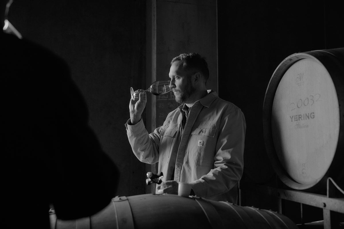 Black and white photo of a winemaker tasting wine beside a large oak barrel in the cellar