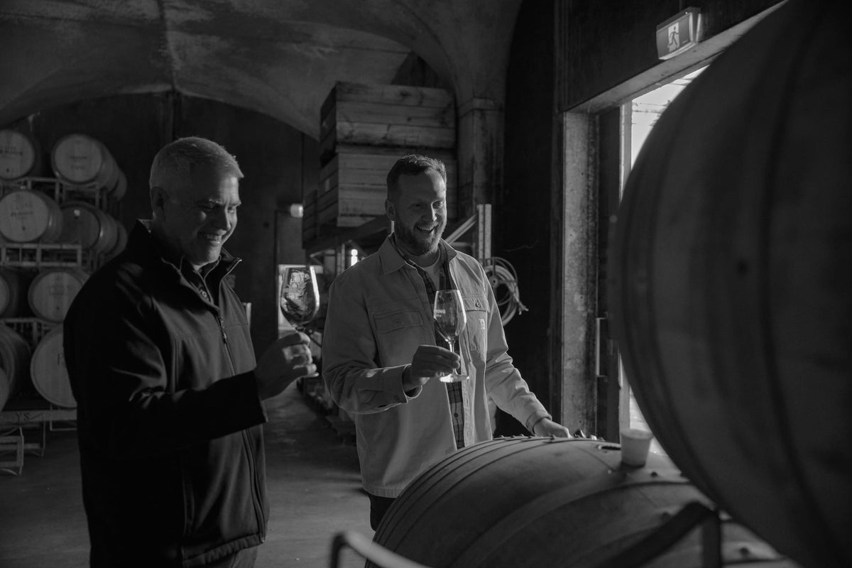 Black and white photo of two men tasting wine among barrels in the cellar