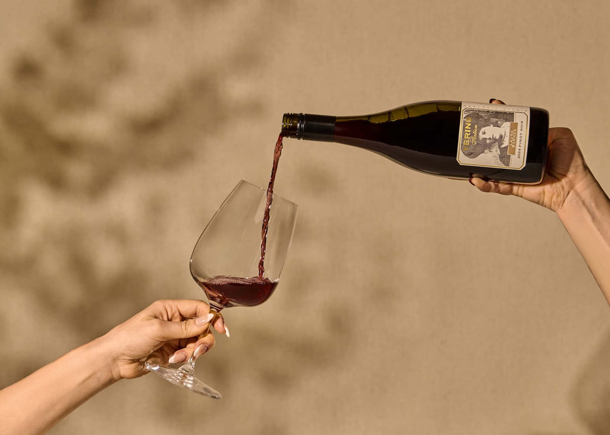 Hands pouring red wine from a bottle into a glass, with a portrait illustration on the label