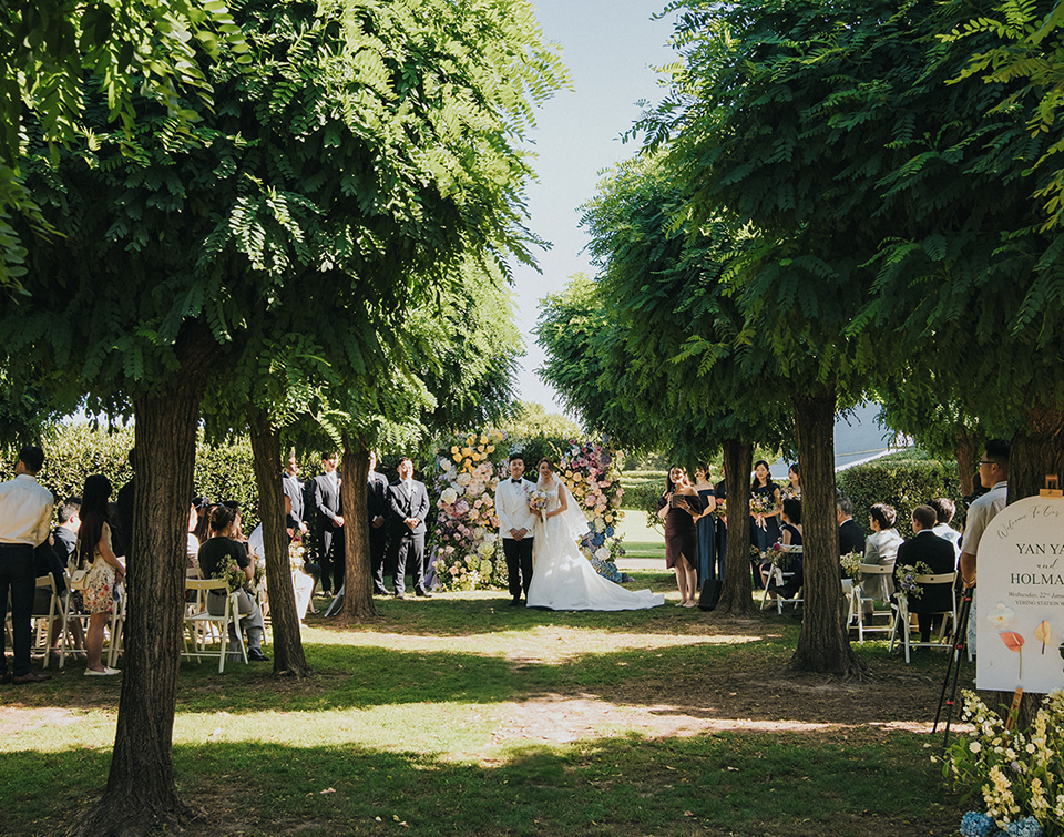 Outdoor wedding ceremony under a tree-lined avenue with seated guests and a floral arch