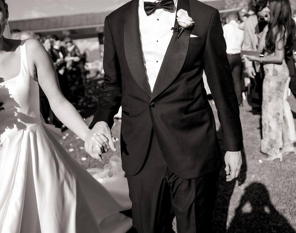 Black and white photo of a bride and groom holding hands while walking past guests