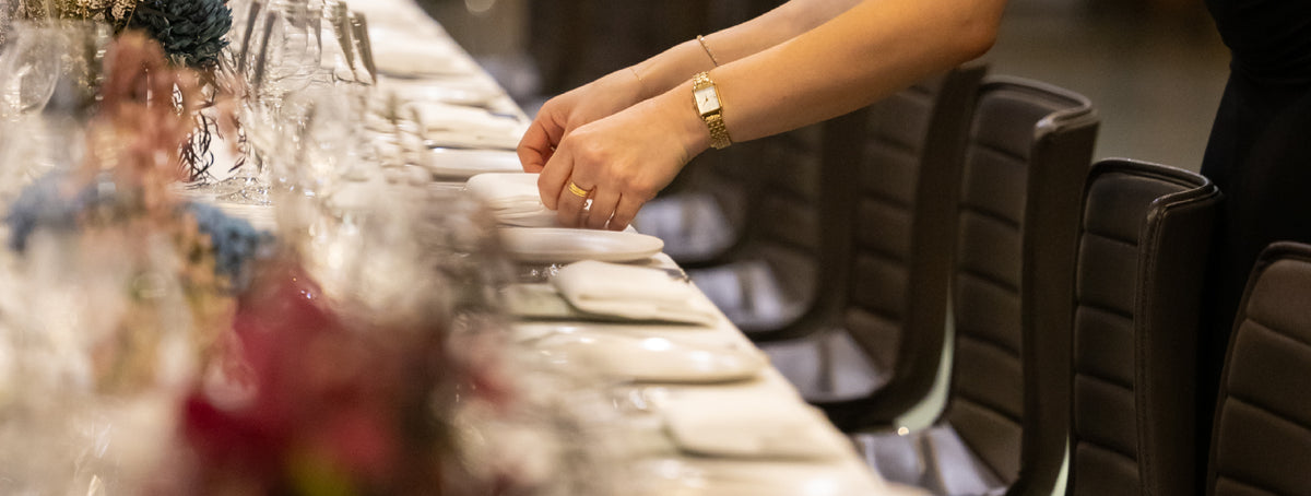 Hands arranging place settings on a long banquet table decorated with flowers