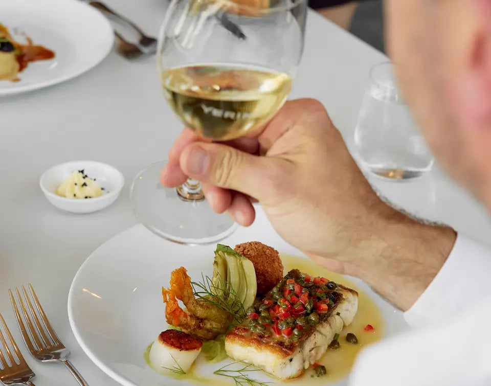 Diner holding a glass of white wine beside a plated seafood dish at a restaurant table