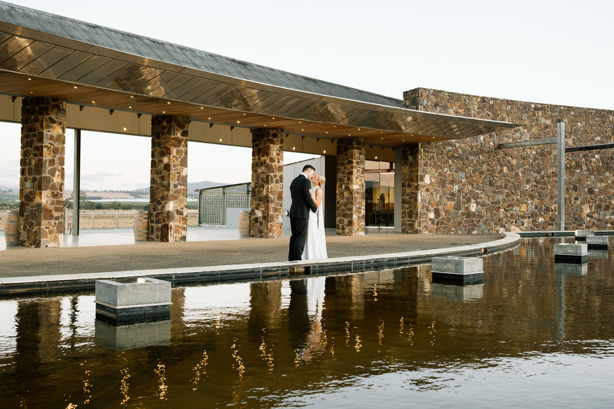 Bride and groom embracing beside a stone building overlooking a reflection pool at dusk