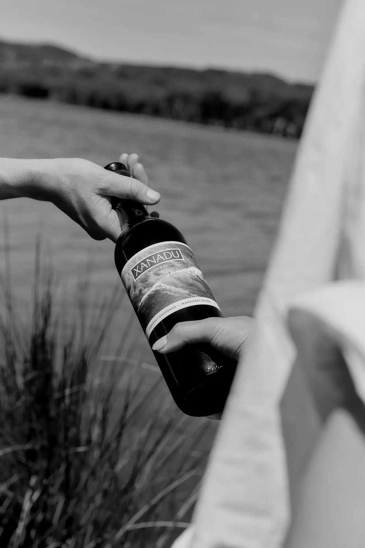Black and white photo of a hand holding a Xanadu wine bottle beside a lake