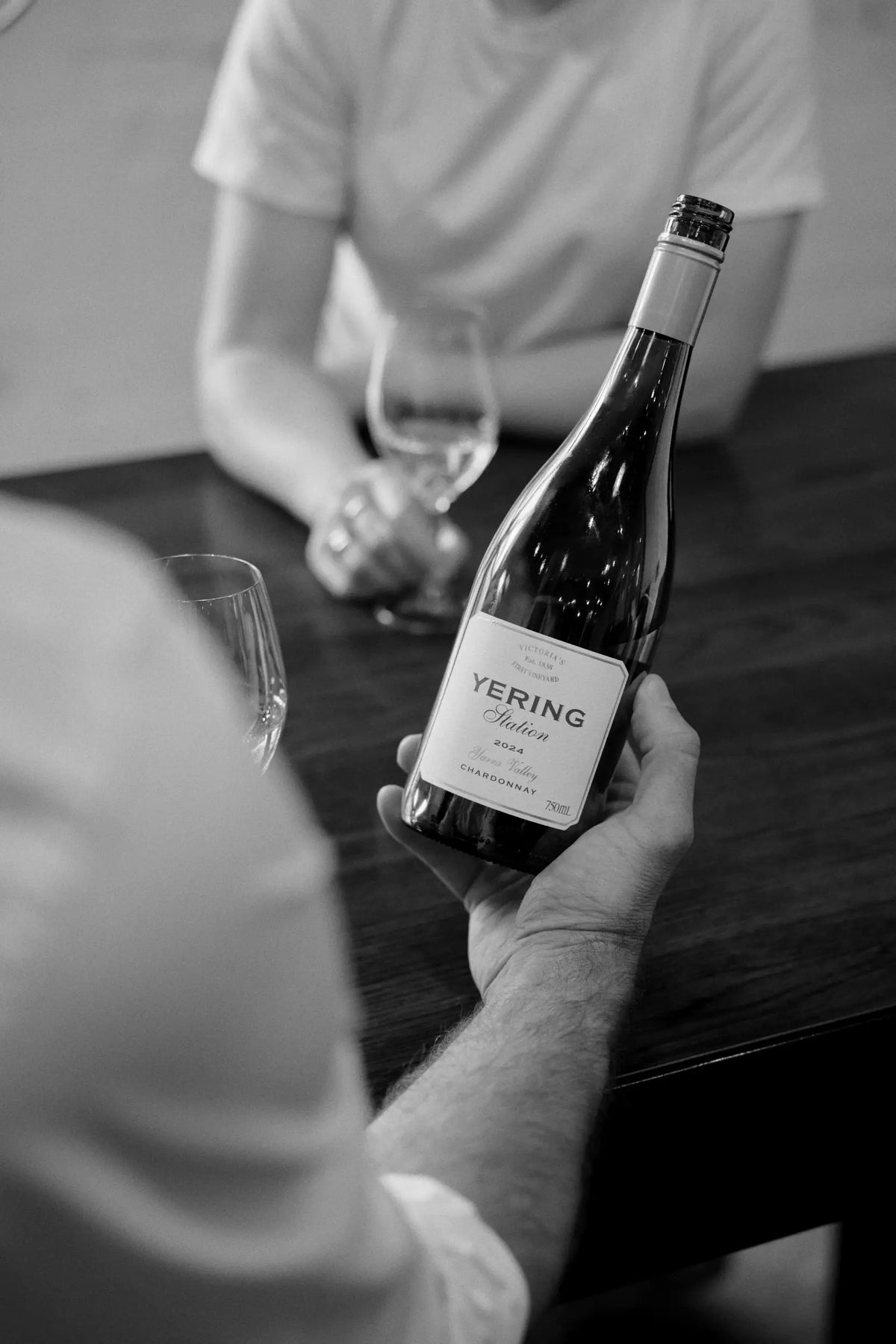 Black and white photo of a hand holding a bottle of Yering Station Chardonnay at a table